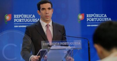 A Portuguese government official speaking at an official press conference behind a podium labeled “República Portuguesa.”
