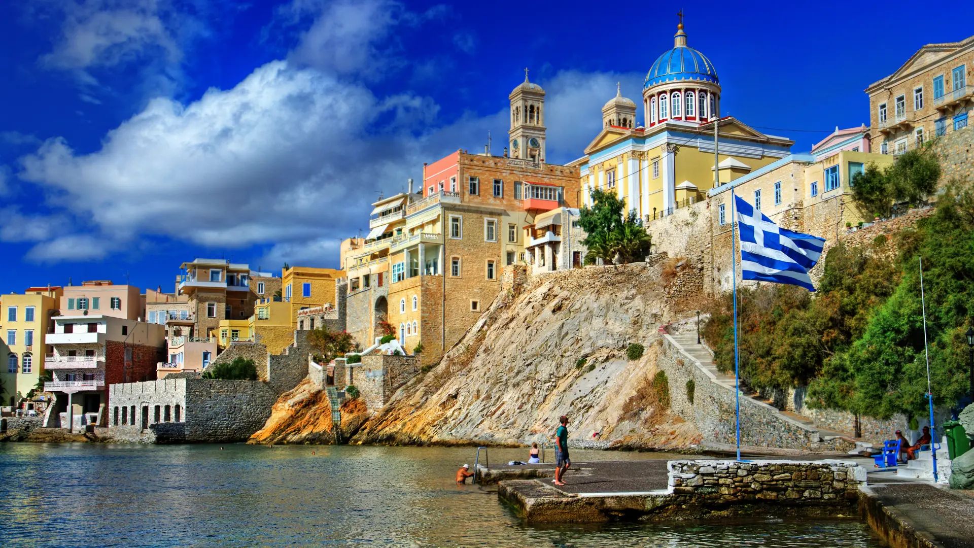 A coastal Greek village built on a rocky cliffside, showcasing historic stone buildings, a blue-domed church, and the Greek flag flying over a stone dock with clear blue water below. (Greek Golden Visa Citizenship)