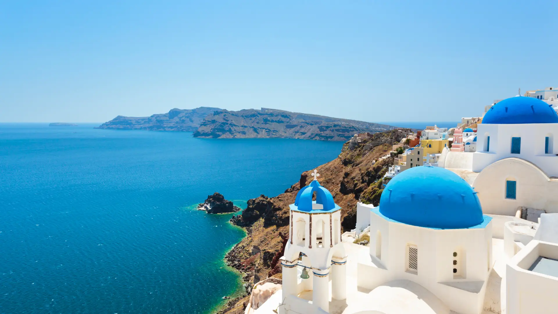 High-angle shot of white-washed buildings and iconic blue-domed churches in Oia, Santorini, set against volcanic cliffs and clear turquoise sea under a bright blue sky. (Greek Golden Visa Citizenship)